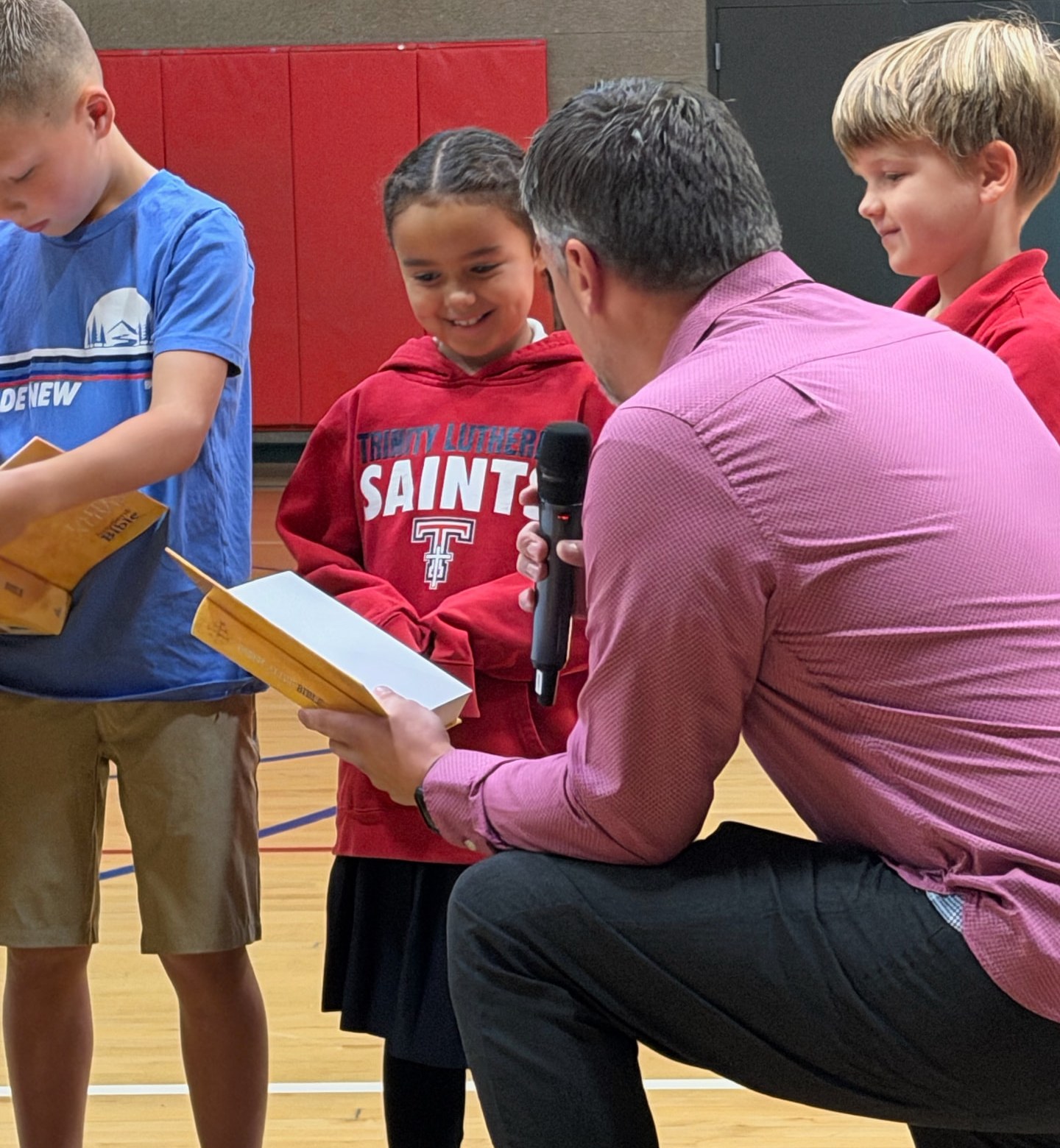 A Trinity leader kneeling with a microphone as a smiling student in a Saints hoodie receives her Bible in the gym