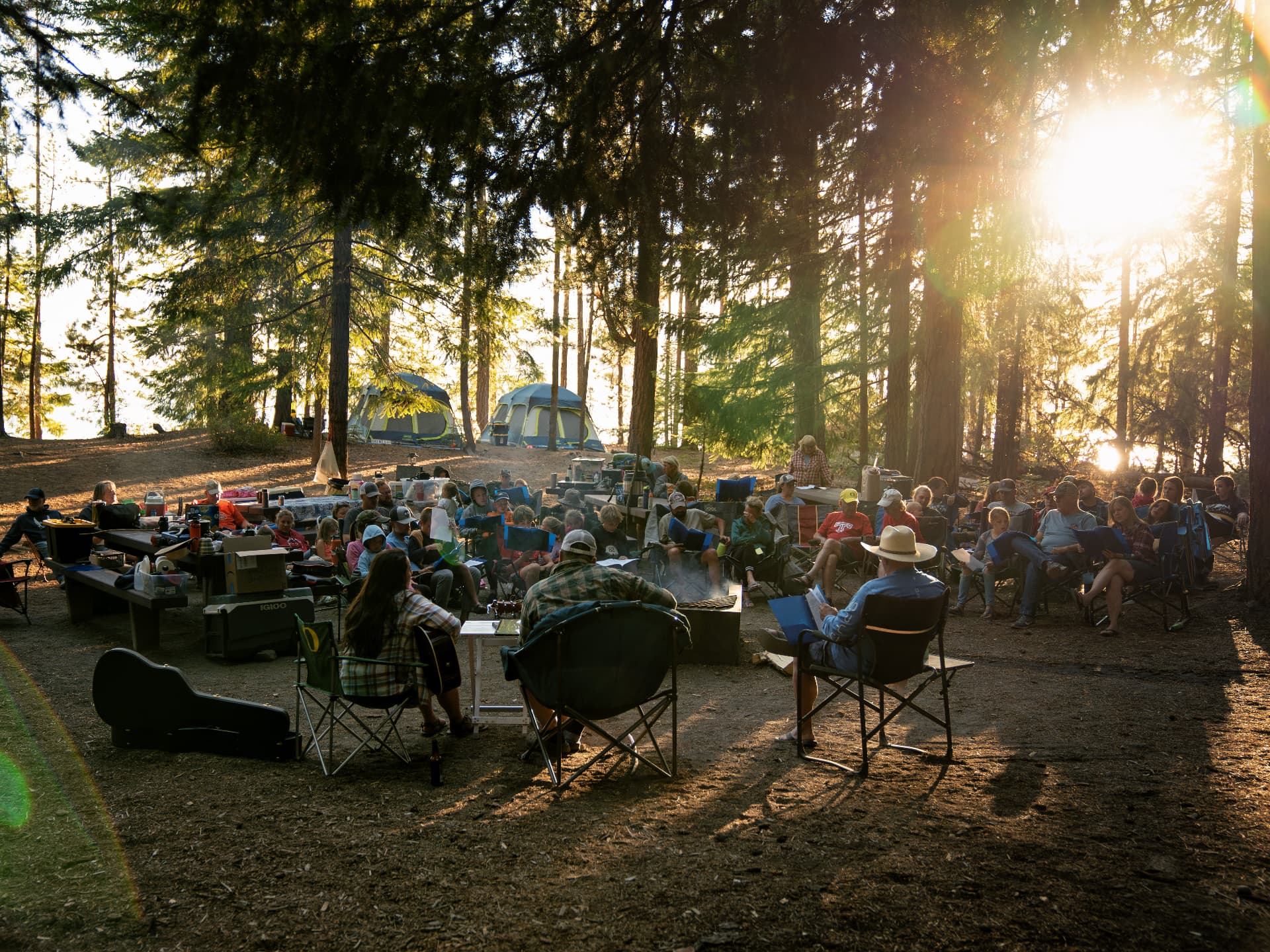 Trinity families gathered in a circle of camp chairs under the pines at Family Camp, singing with a guitar